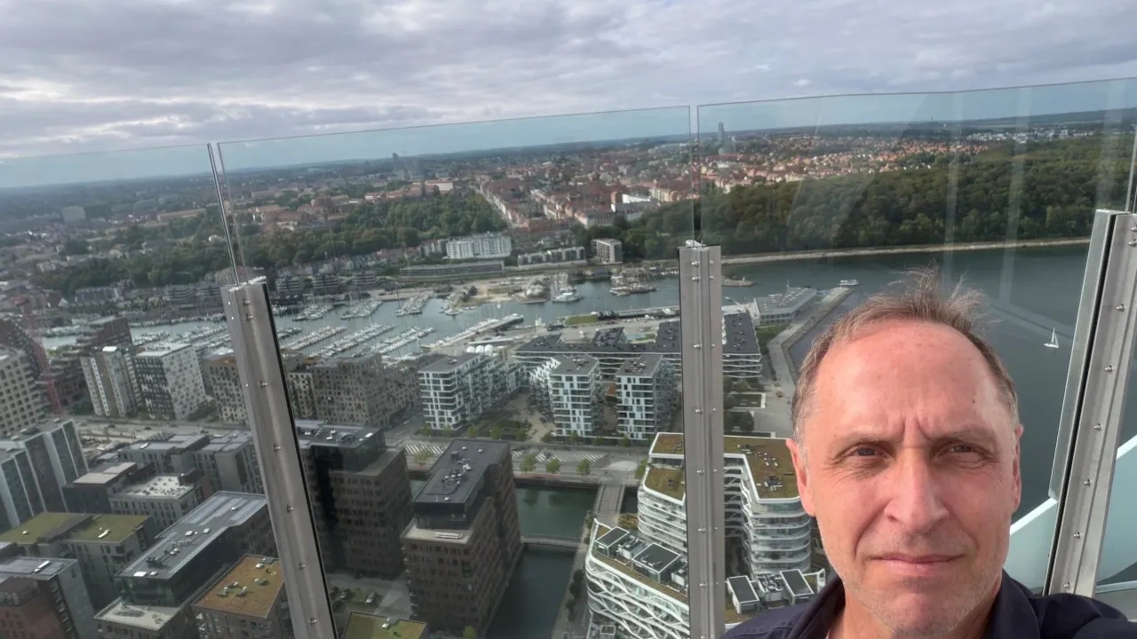 David Erichsen på en udsigtsplatform med glasværn og panoramaudsigt over Københavns havn, moderne bygninger, lystbådehavn og grønne områder.
